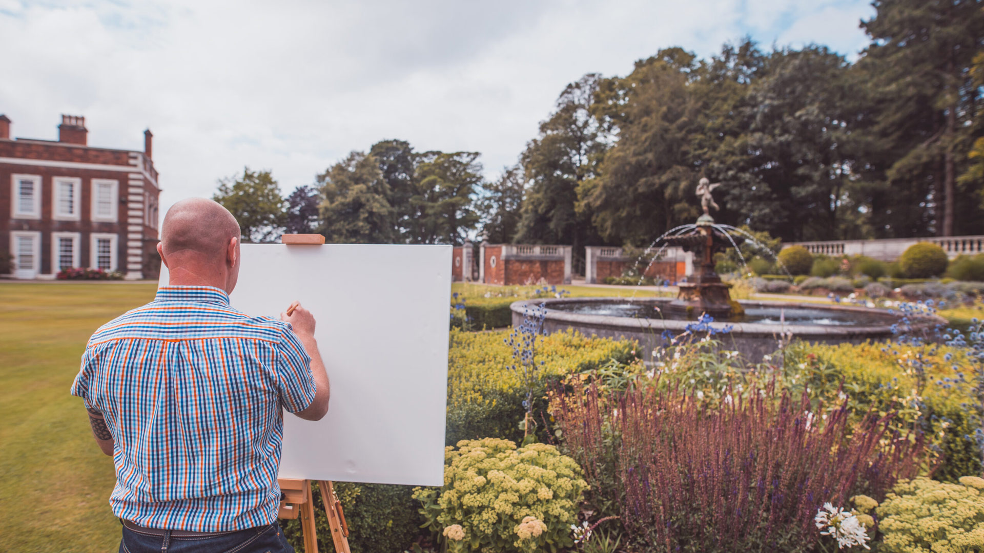 The back of a man with an easel and standing in beautiful gardens of Knowsley Hall with a running fountain ready to paint a picture